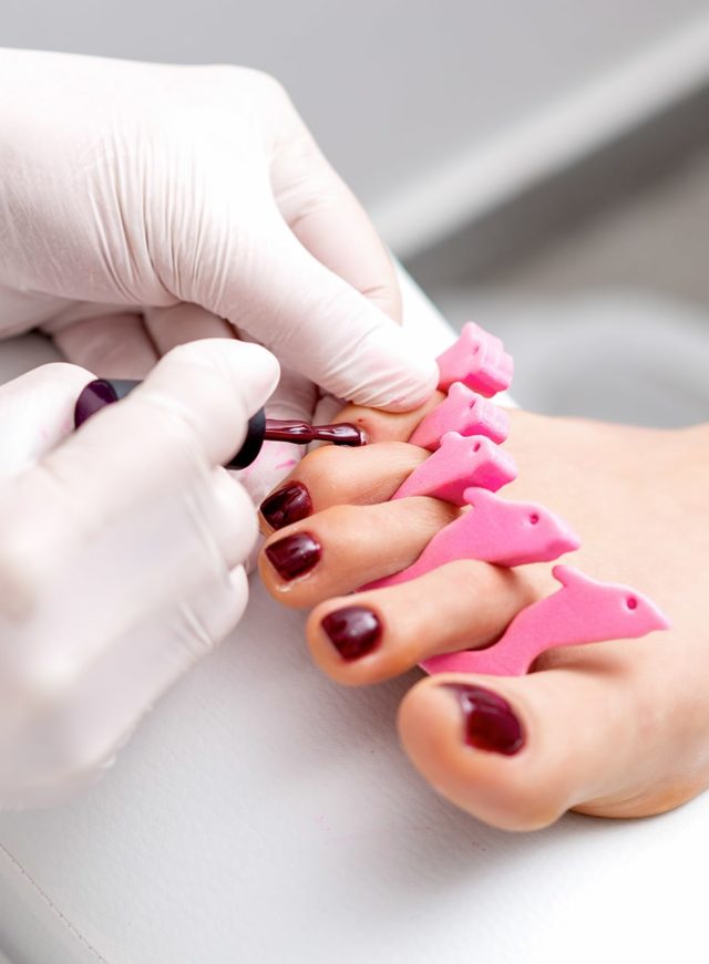 manicure-master-is-painting-female-toenails-with-maroon-nail-polish-by-brush-wearing-white-gloves
